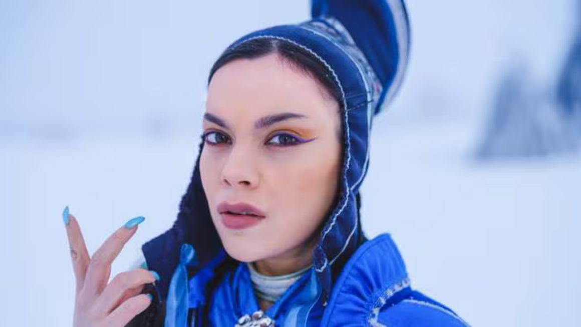 Close-up of a woman wearing a blue outfit and headpiece with blue and yellow eye makeup, posing outdoors in a snowy landscape"