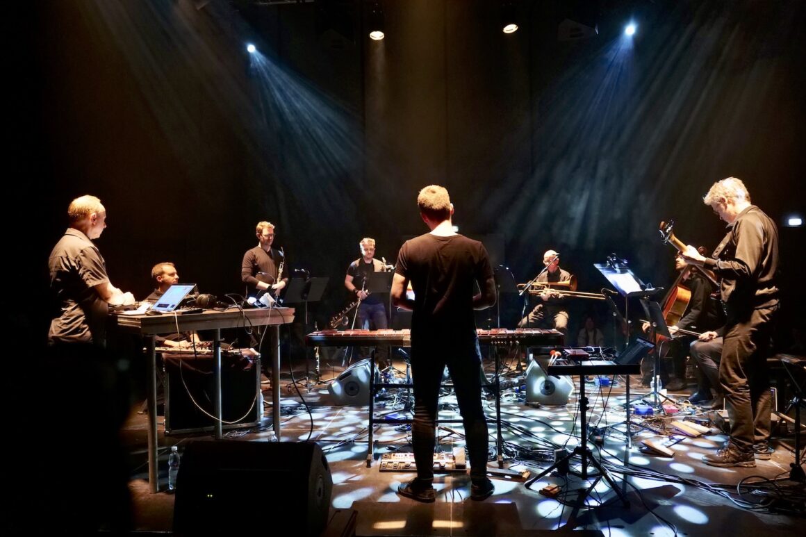 A series of figures, mostly men, standing on stage, playing an array of instruments, with the overhead lights focusiing on the floor in a dramatic way