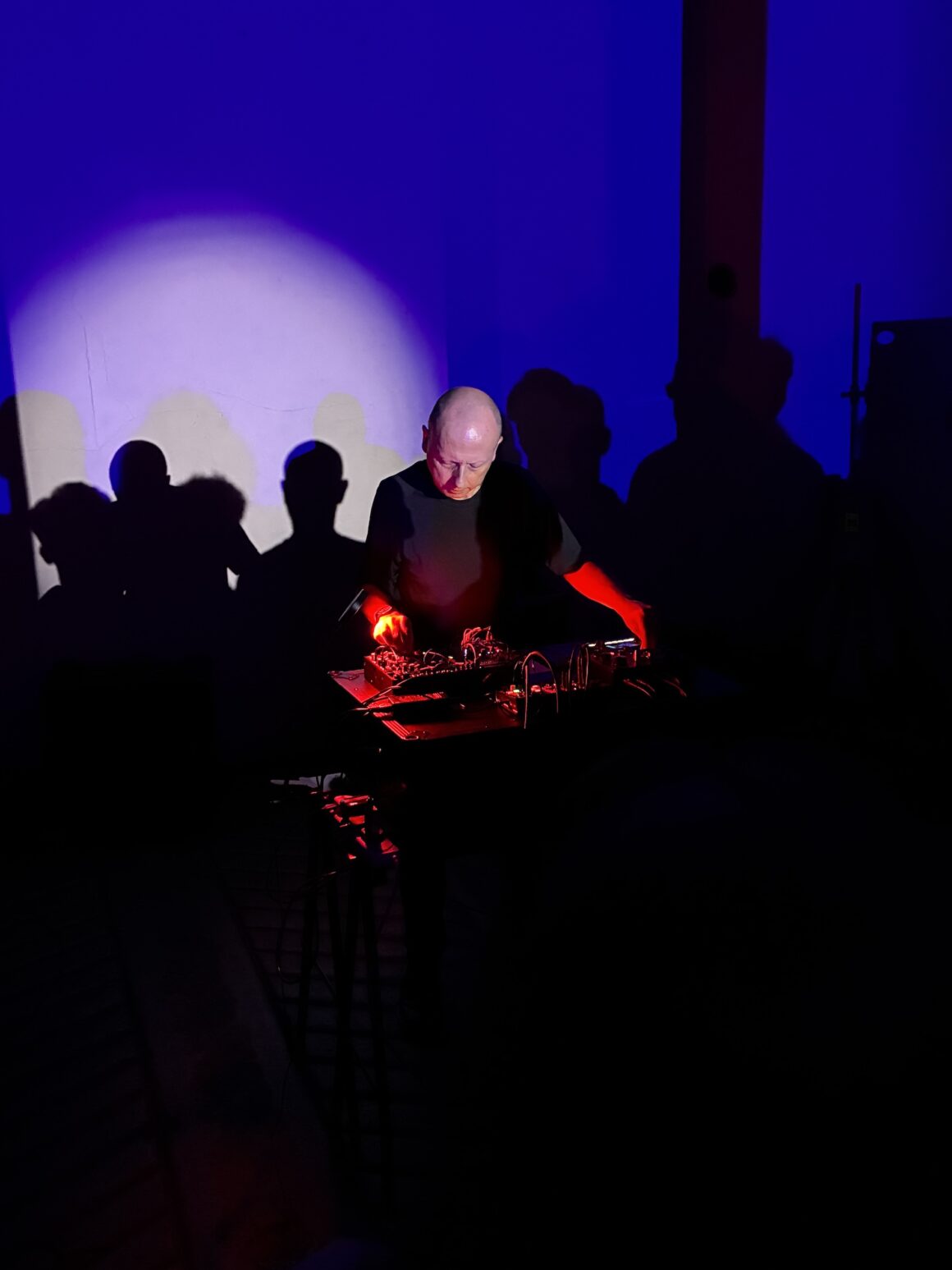 Electronic music performer illuminated by red and blue lights, standing behind a table with music equipment in a dark room with shadows of an audience on the wall.
