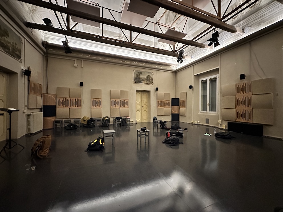 A large empty room, in a theatre, the ceiling lights reflecting on the floor. A few items, bags and chairs, sit on the floor