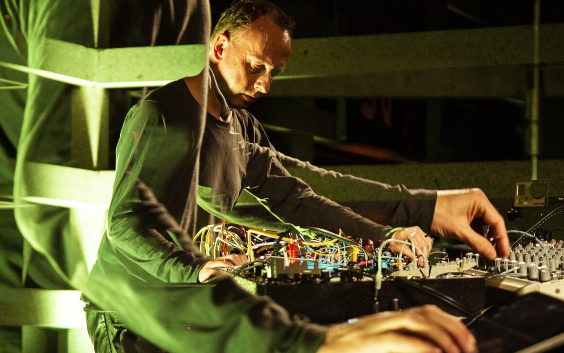 Colourful image of a white man standing over a table of electronic musical equipment, collaged together with a close-up of his hands turning knobs