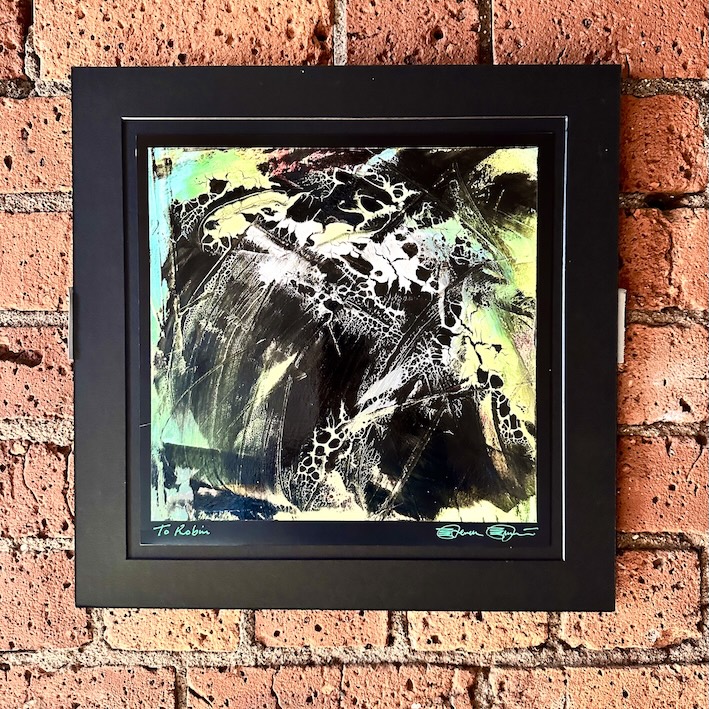 A very colourful abstract painting hung on a brick wall. The image is framed with a thick black frame and has green and white ink washed across it