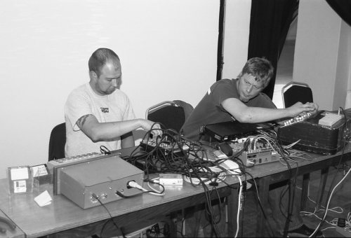 Black and white image of two men sitting at table covered in electronic music gear