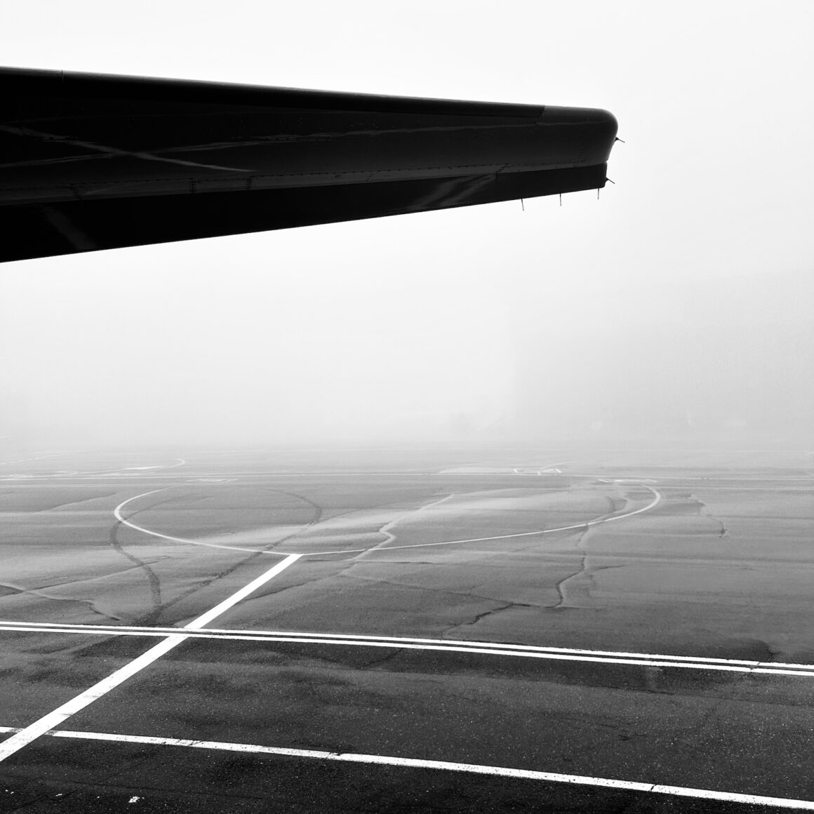 Black and white image of the wing of a plane seen from the side at an aiport, with the ground just below
