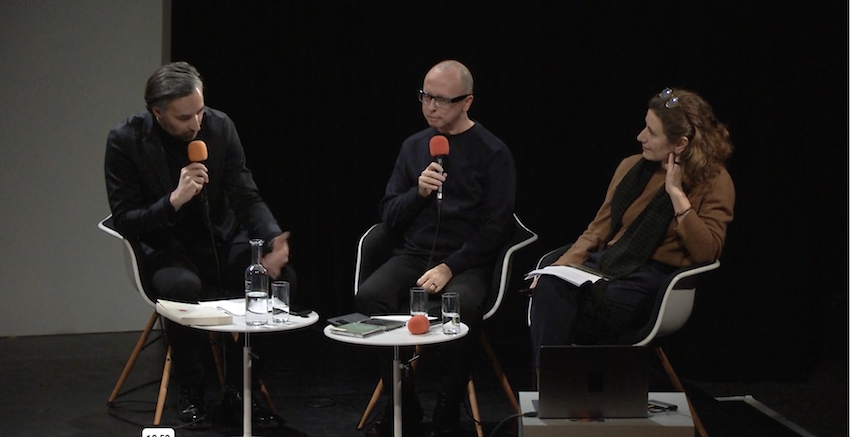 Three people, two men and one woman, sit at a table holding microphones which look like lollipops. They are in conversation sitting in front of a small white table