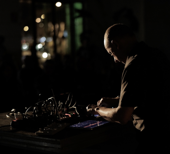 Man operating electronic music equipment in a dark room with dim lighting and blurred background.