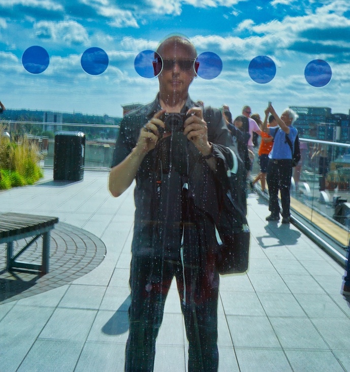 Very blue coloured image of a man photographing himself in the reflection of a window, with six dark blue dots across to top of the image. The man is white, with cropped hair, wearing black sunglasses and wearing dark clothes holding a camera pointing towards the centre of the image
