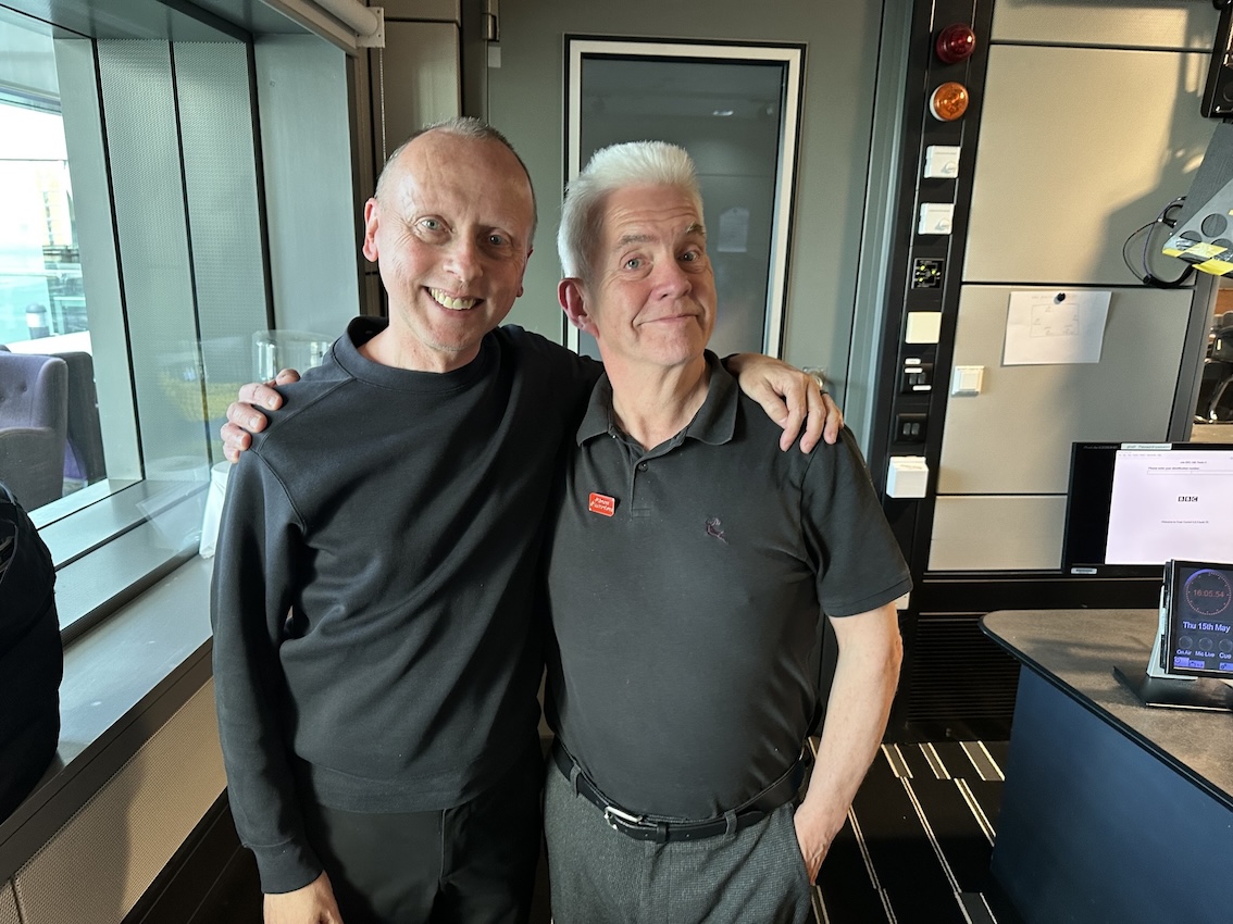 Two older men stand shoulder to shoulder, proudly smiling at the camera. Both are dressed in black and standing in a studio at the BBC