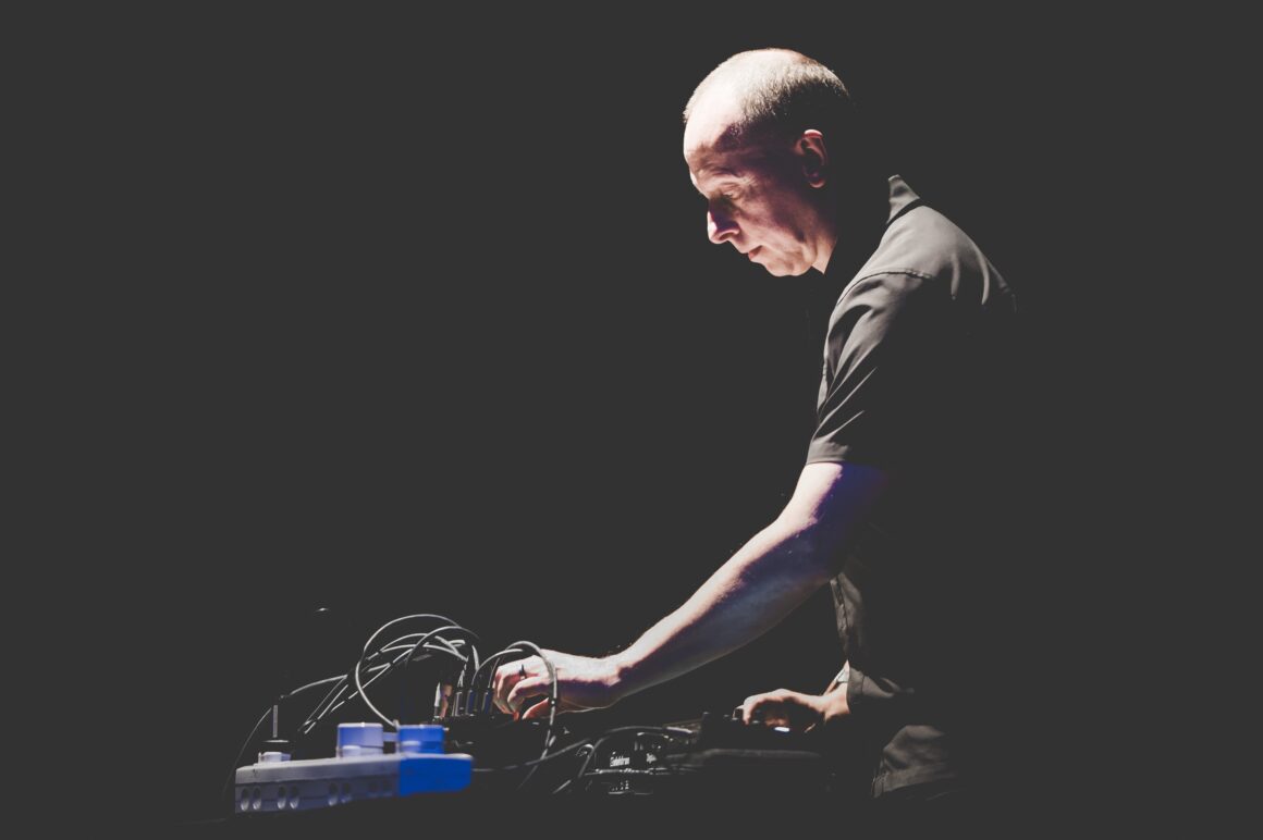 Man standing behind electronic music equipment and adjusting it, against a dark background.