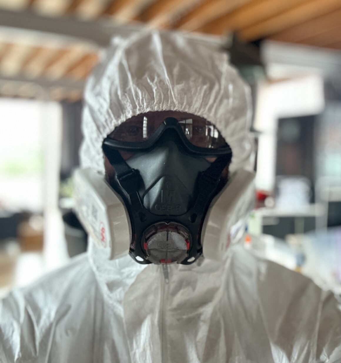 A man wears a PPE suit, all white, with googles and black industrial mask with ventilator, standing inside a residential house, close up.