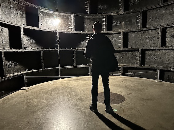 A man seen from behind stands in an industrial space with a bright bulb in front of him. You cannot see his face