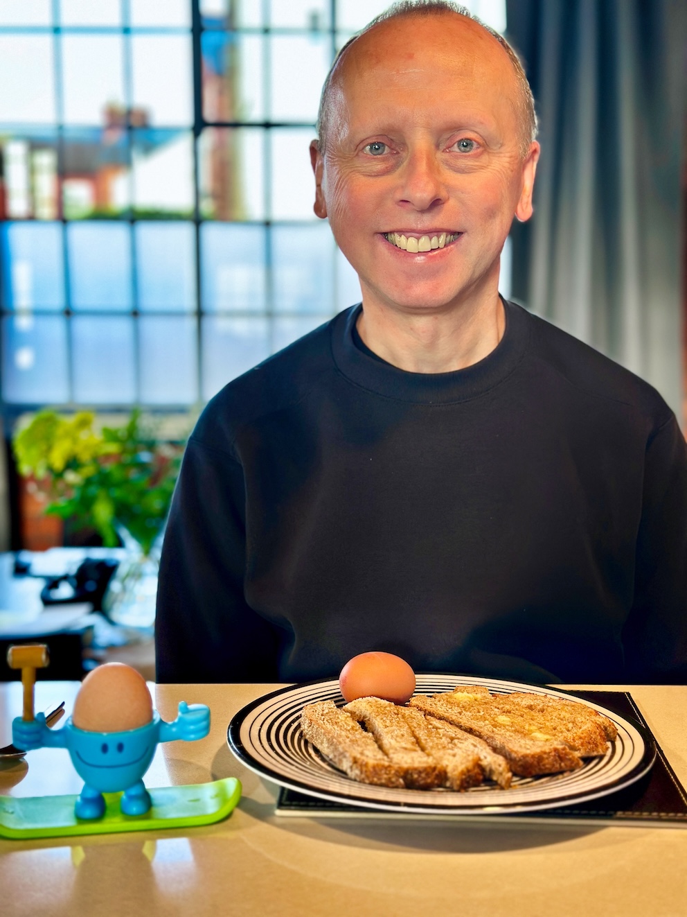 A handsome white man with short hair sits at a table, smiling happily directly at the camera, dressedin a black top. Before him sits toast and an egg, in a celebratory happy manner