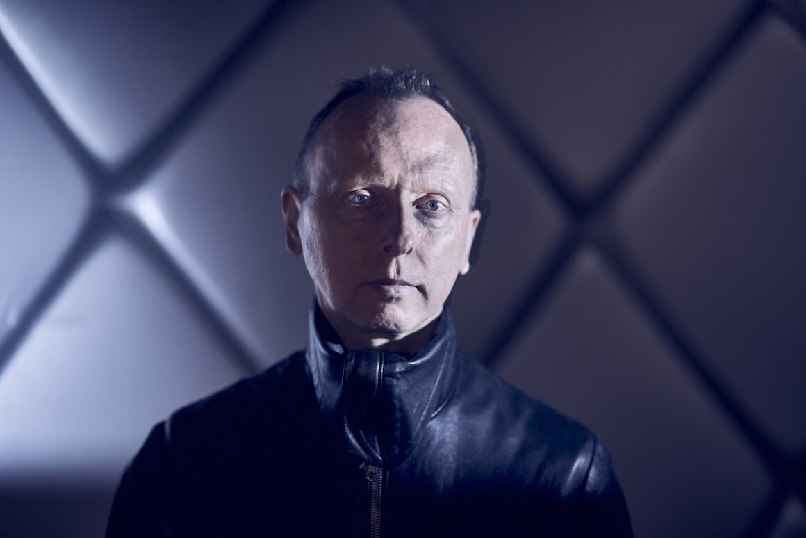 colour portrait photograph of older white man with short hair, staring directly at the camera, only his shoulders visible. He is wearing a shiny black leather jacket, standing before an illuminated background of patterns