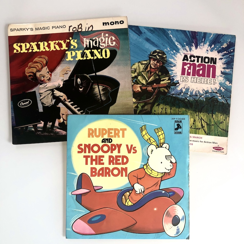 3 x 7" singles on a white surface, with colourful sleeves featuring illustrations. They are Sparky's Magic Piano, Action Man is Here and Rupert and Snoopy