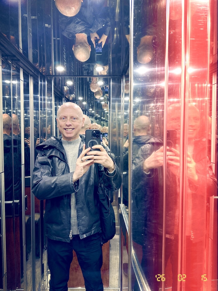 Man smiling, wearing a black leather jacket and gray sweater taking a selfie with a smartphone inside an elevator with mirrored walls and ceiling.