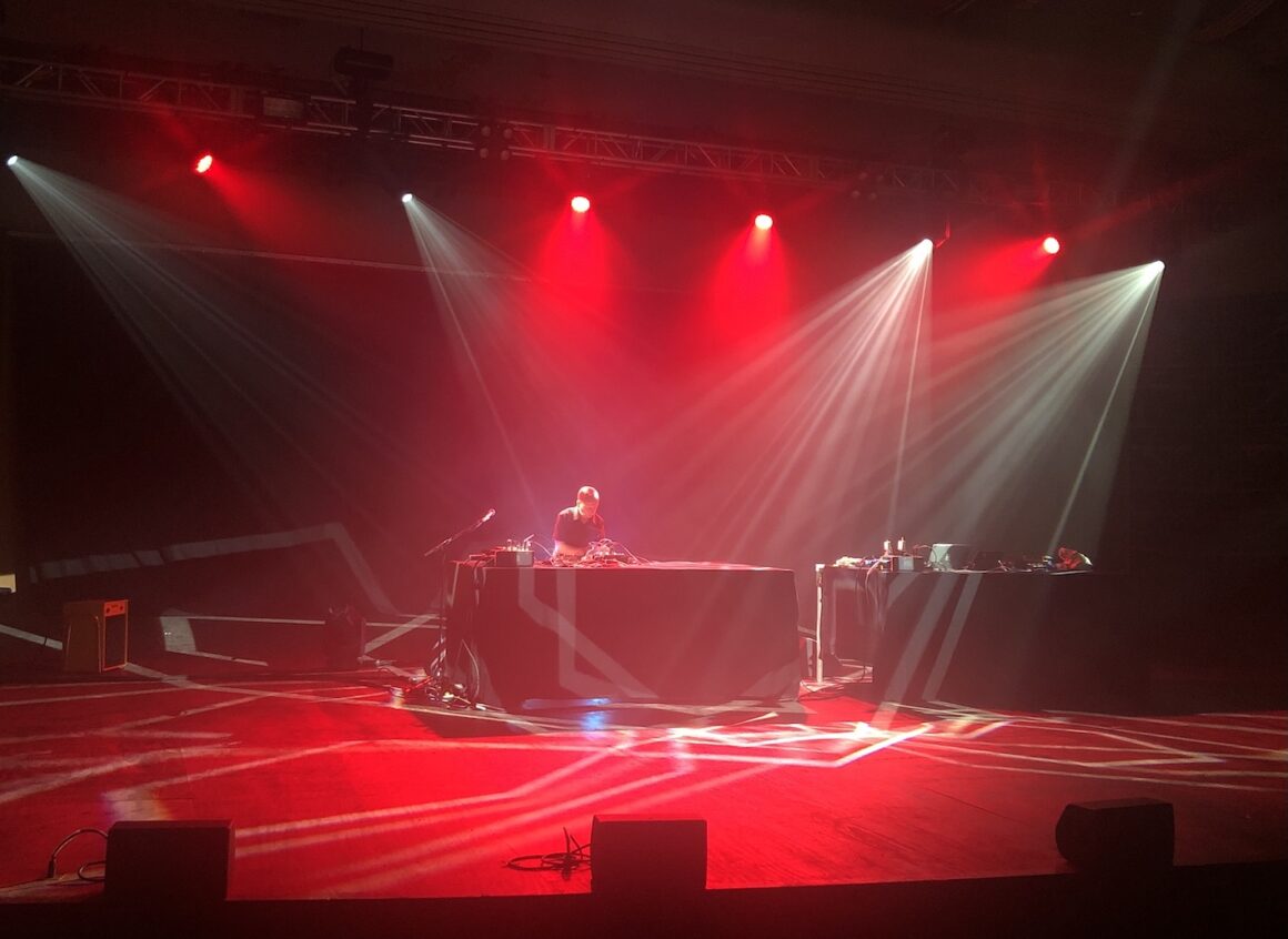 A dramatic image of a man on stage wearing a black short sleeve shirt, standing behind a large table, with bright lights shining on him from above, creating patterns across the stage floor in front