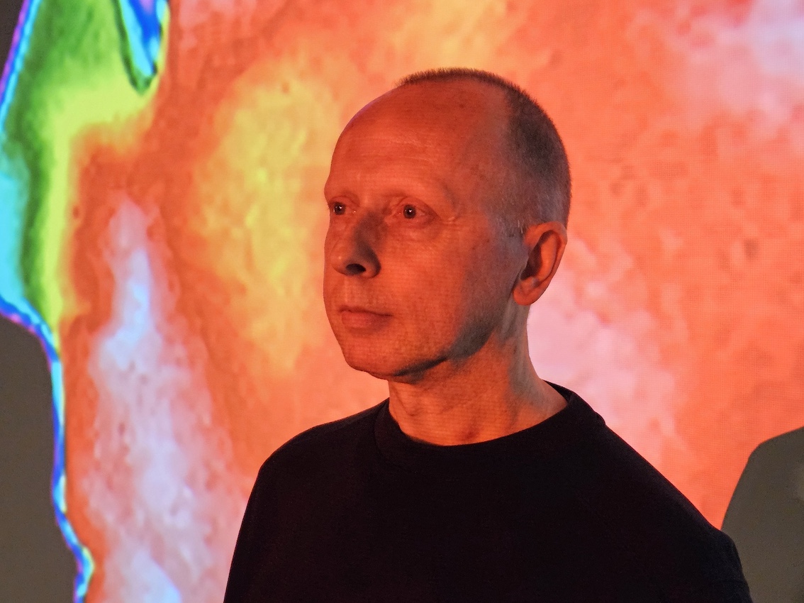 A white man can be seen from the shoulders up, dressed in a black top. He has short cropped hair and is staring off into the distance. Behind him a very abstract colourful image is displayed, making the entire image red and glowing