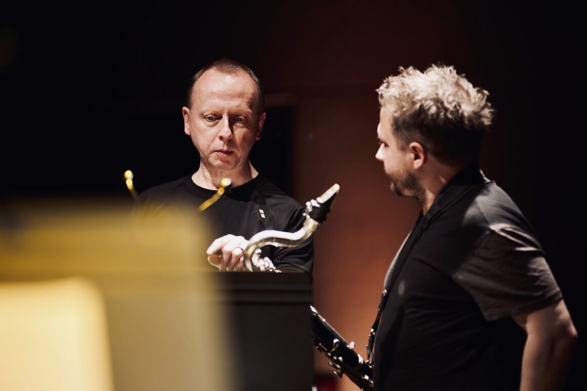 Two men stand together one holding a bass clarinet as they talk deep in conversation