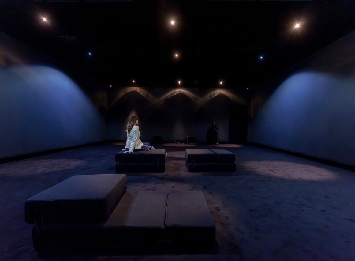 Dark room interior with blue lighting effects on walls, a woman sitting on a cushioned bench in the middle, and several cushioned benches arranged on the floor.