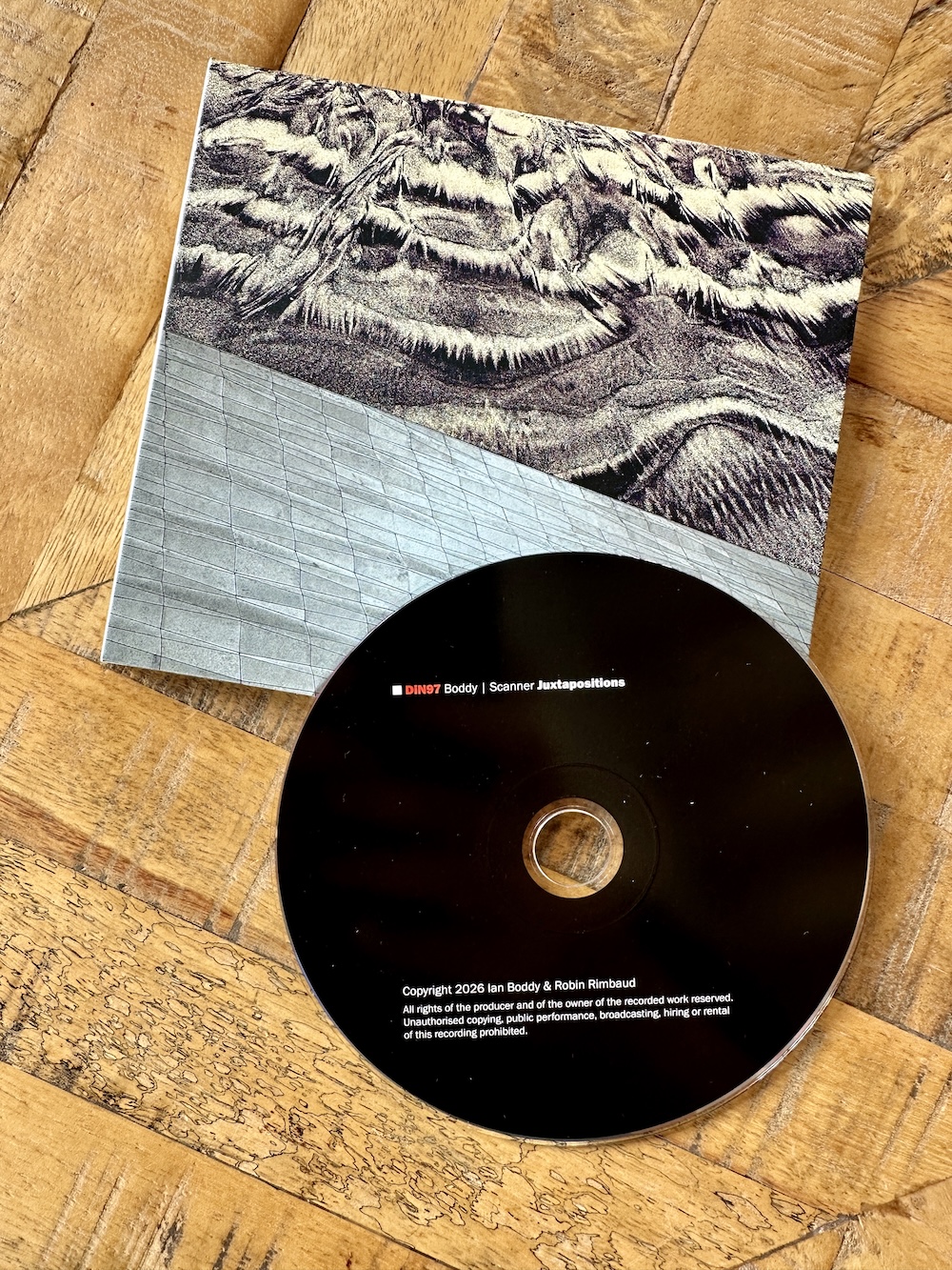 A CD titled "DIN97 Boddy | Scanner Juxtapositions" along with its booklet featuring an abstract textured design, placed on a wooden surface.