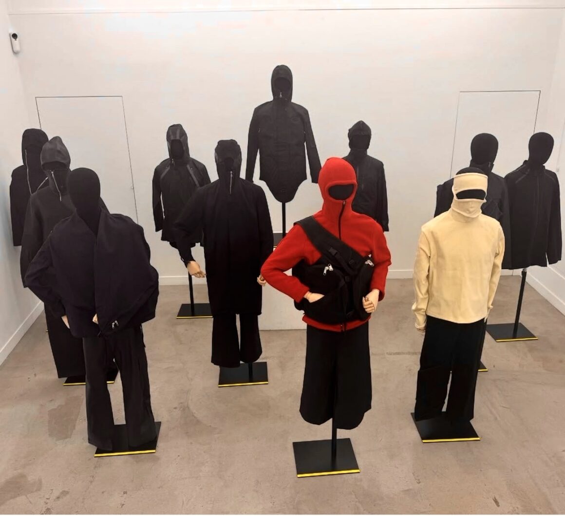 Display of mannequins dressed in black clothing, with two mannequins in the front wearing a red outfit and a beige outfit with full-face hoods, standing in a minimalist gallery space