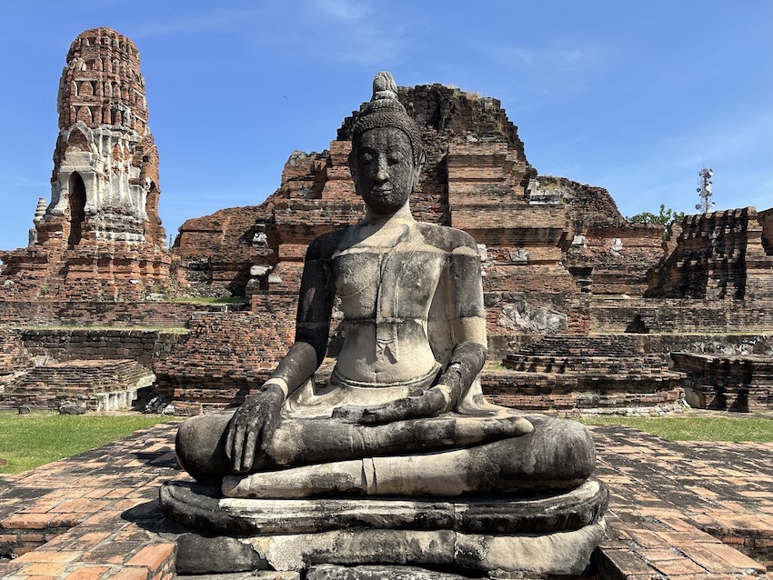 Stone Buddha statue sitting in lotus position in front of old brick ruins under a blue sky.