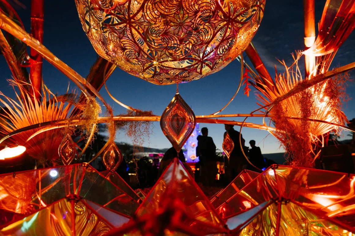 A colourful image, red and golden, where you are seemingly looking through a sculpture in an outdoor music festival at night and can see a few people in the crowd in front of you are but they just dark shadows
