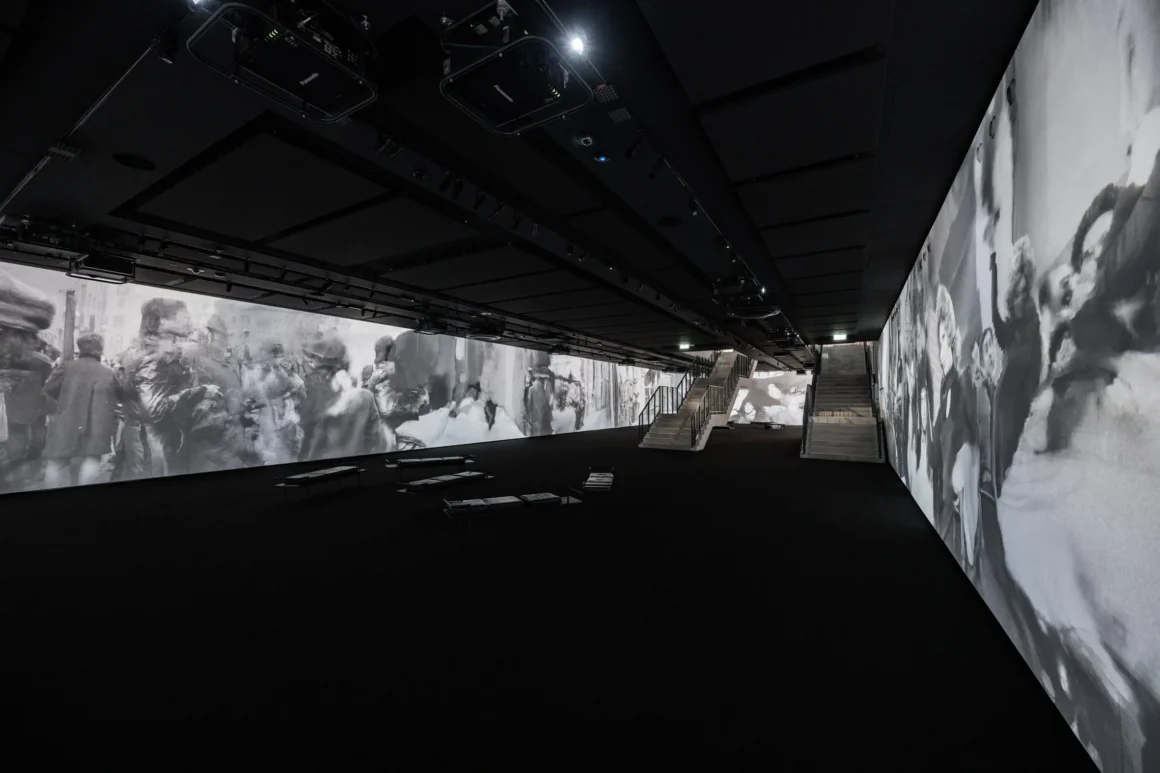 Black and white photograph of inside a large gallery space with large video projections on the surrounding walls